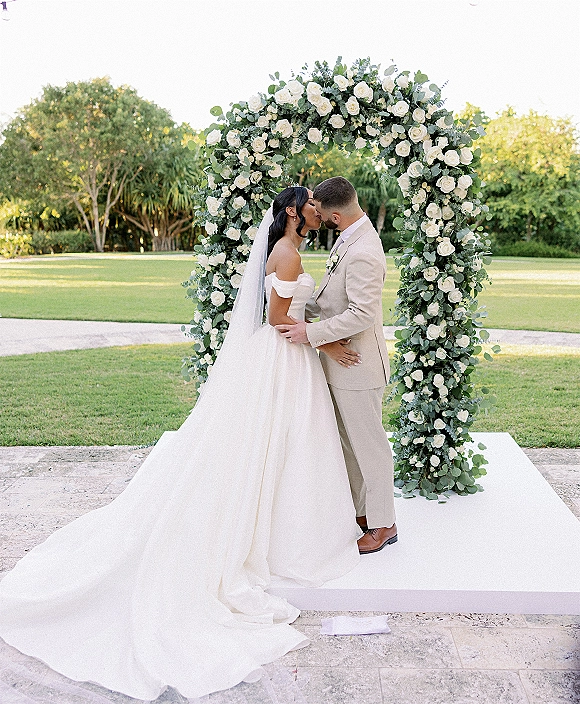 Wedding kiss as bride and groom embrace under a white rose and greenery floral arch, veil and off-shoulder dress on garden patio stage