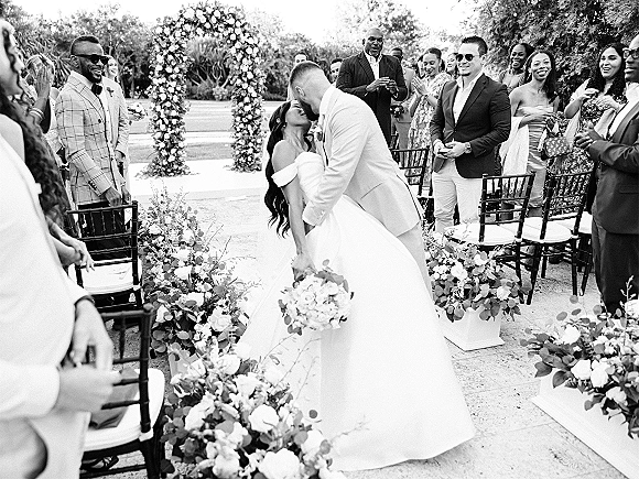 Wedding kiss as the couple dips under a floral arch, bride in an off the shoulder gown holding a bouquet along a garden ceremony aisle