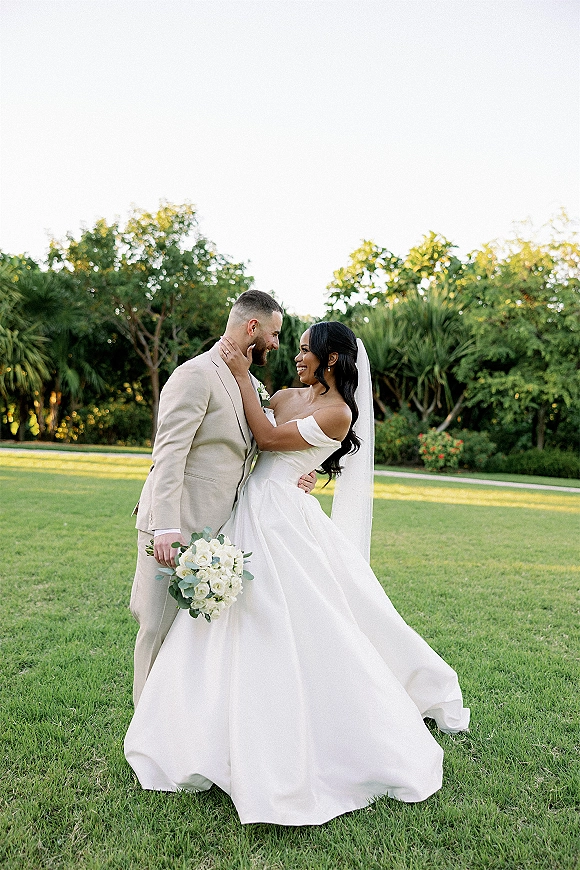 Couple portrait outdoors with bride and groom embrace, bride in strapless dress and long veil holding a white rose bouquet on a lawn