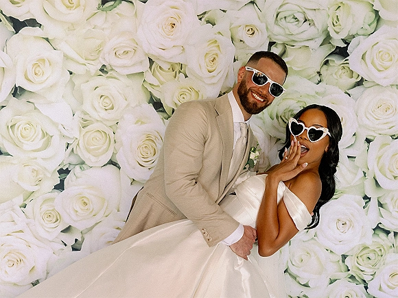 Couple portrait of bride and groom sunglasses as he dips her in a strapless gown and beige suit before a white rose floral wall backdrop