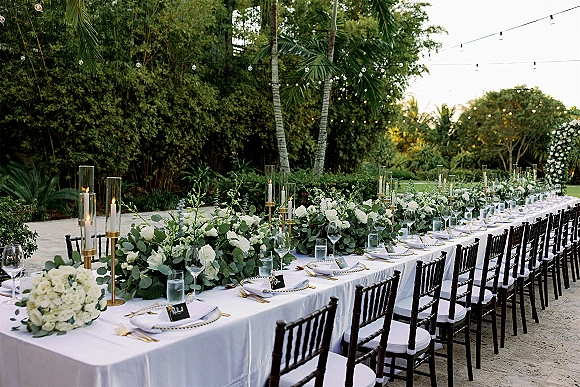 Reception tablescape on a long banquet table with white roses, greenery runner, gold candlesticks, and glass candles under string lights in a garden