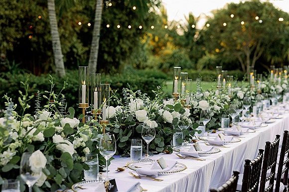 Reception tablescape with outdoor wedding tablescape styling, white rose and greenery garland, gold candlesticks, and glass cylinders under string lights in a garden