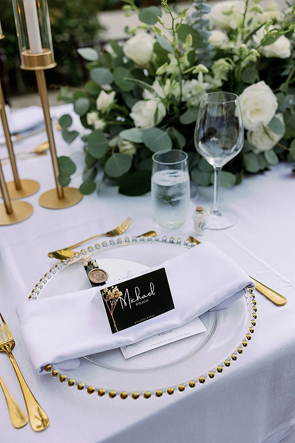 Reception tablescape with a wedding place setting featuring beaded charger, gold flatware, candles, white roses, and eucalyptus in garden light
