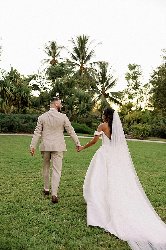Couple portrait of newlyweds holding hands from behind, bride’s long veil trailing on grass, groom in beige suit amid palm trees