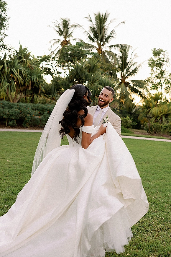 Couple portrait of bride and groom laughing as he lifts her, long cathedral veil blowing on a palm-lined lawn under open sky