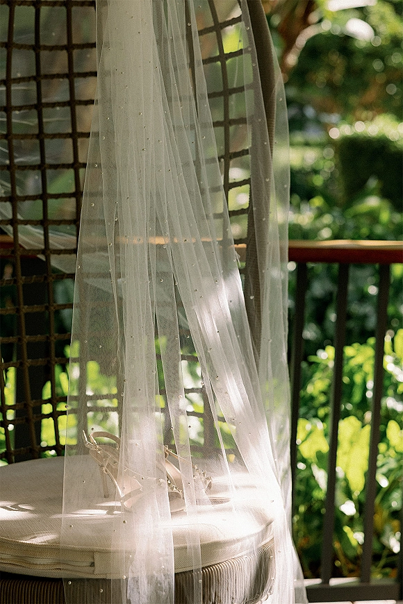 Bridal veil with beaded wedding veil detail draped over a cushioned chair beside bridal shoes, sunlit greenery and lattice behind