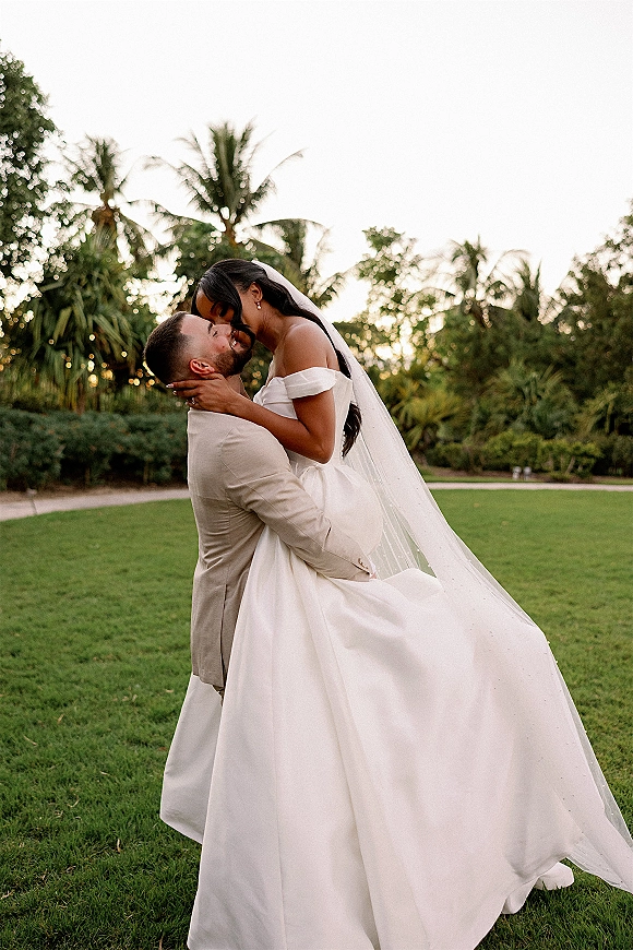 Wedding couple portrait of the bride and groom kiss as he dips her, veil flowing over an off-the-shoulder dress on a palm-lined lawn