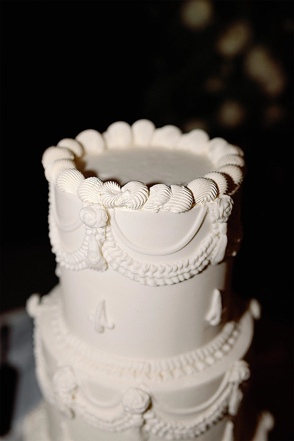 Wedding cake with white wedding cake buttercream swags, rosettes, and scalloped borders on a cake table against dark bokeh lights backdrop