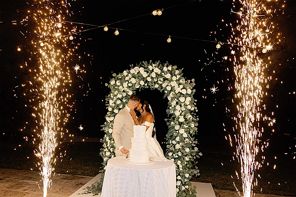 Wedding cake cutting as bride and groom kiss by a tiered white cake under a rose greenery arch, cold spark fountains and string lights at night