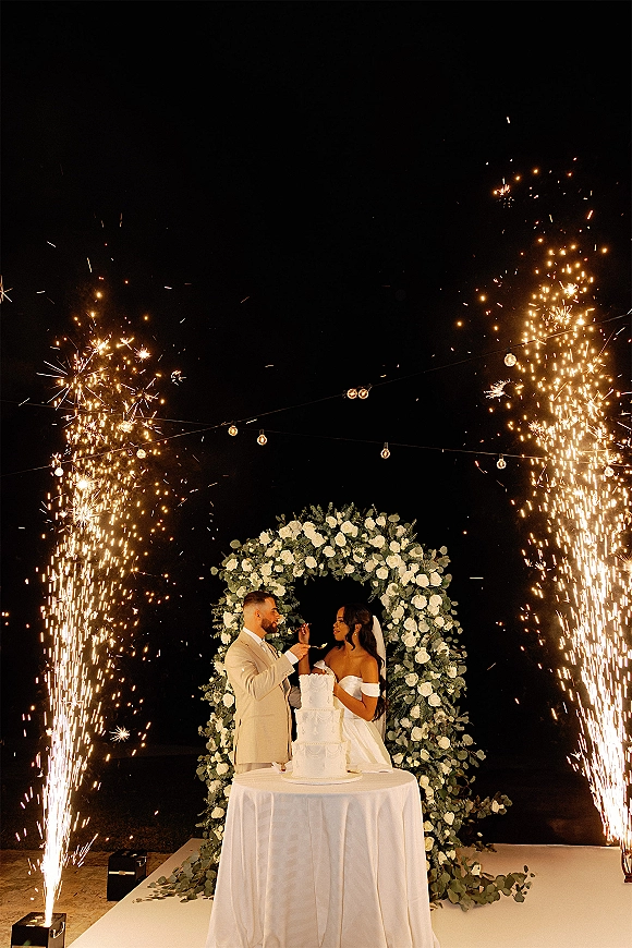 Wedding cake cutting as bride and groom slice a three-tier white cake under string lights, with sparkler fountains and a floral arch at night