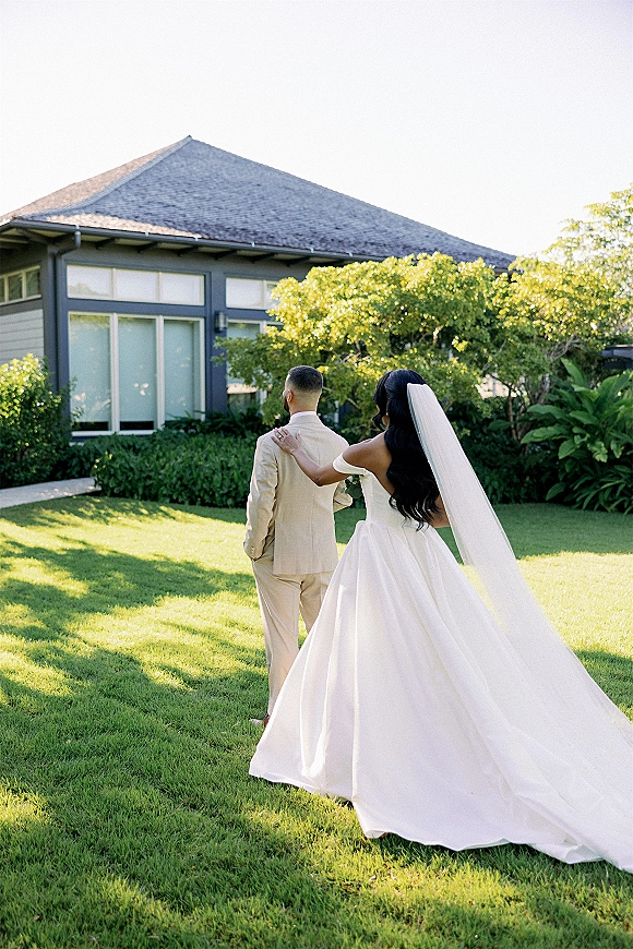 First look moment outdoors as bride taps groom’s shoulder, her long veil and ball gown train flowing on a sunny lawn by a modern venue