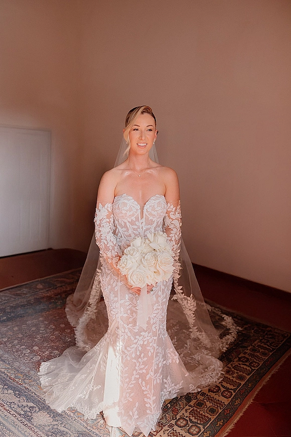 Bridal portrait of a bride in a lace mermaid wedding dress with cathedral veil, holding a white rose bouquet in an indoor doorway setting