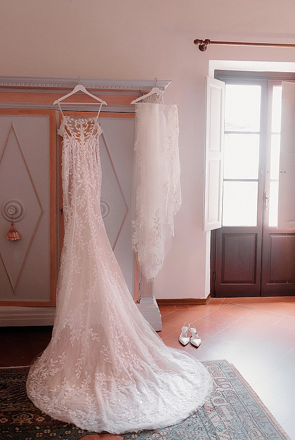 Wedding dress on hanger, lace wedding dress with veil and bridal shoes on a rug by an open window with shutters and armoire