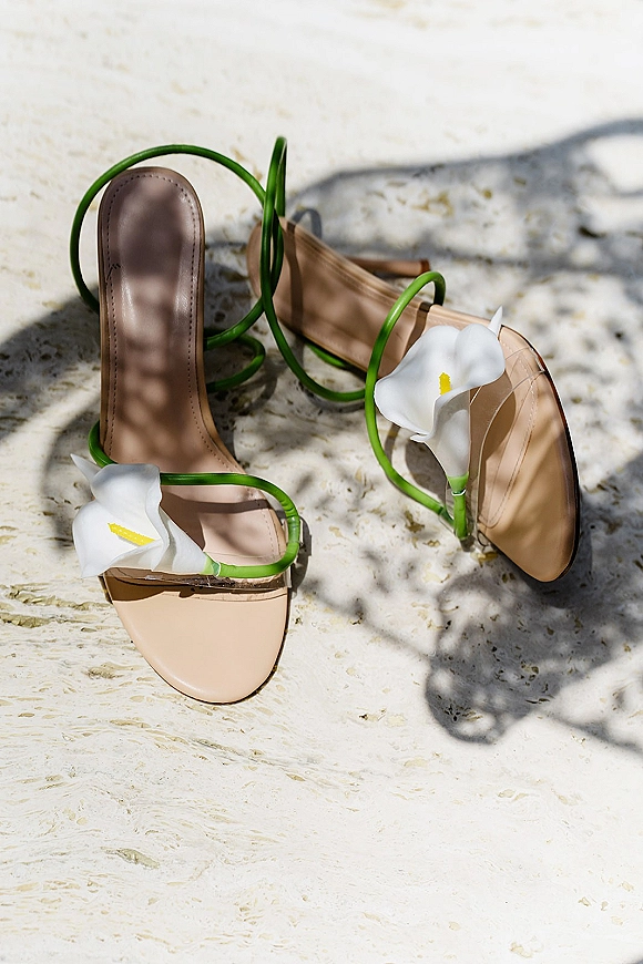 Bridal shoes in a nude flat lay with calla lily accents and green straps on a light stone surface with soft plant shadows