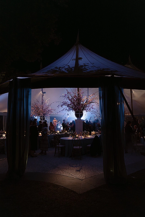 Wedding reception tent with sailcloth drapes, round tables, floral centerpieces and candlelight, as guests gather by the dance floor under night sky