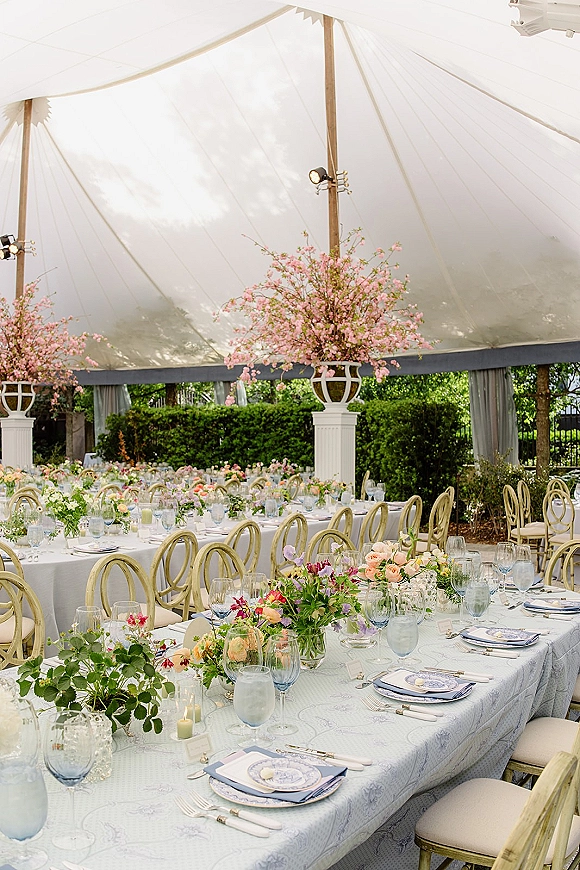 Reception tablescape with outdoor tent reception decor, pastel floral centerpieces and pink branches, blue goblets, pillar candles, and place cards under a white canopy