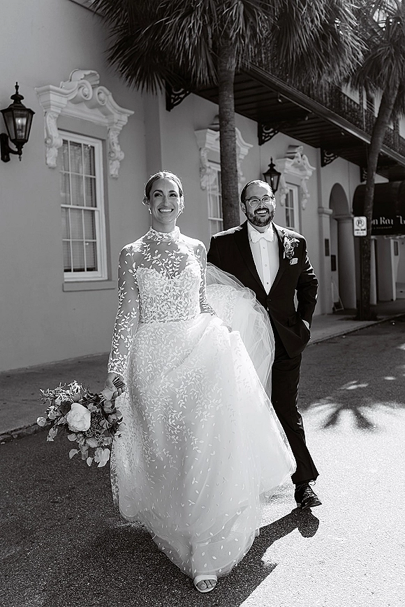 Couple portrait in black and white, bride in long-sleeve lace dress with bouquet and veil walking beside groom in tuxedo on palm-lined street