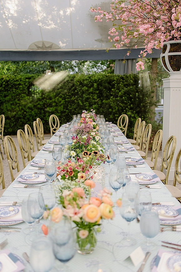 Reception tablescape on a long banquet table with pastel floral centerpieces, blue stemware accents, and place cards under a draped tent canopy