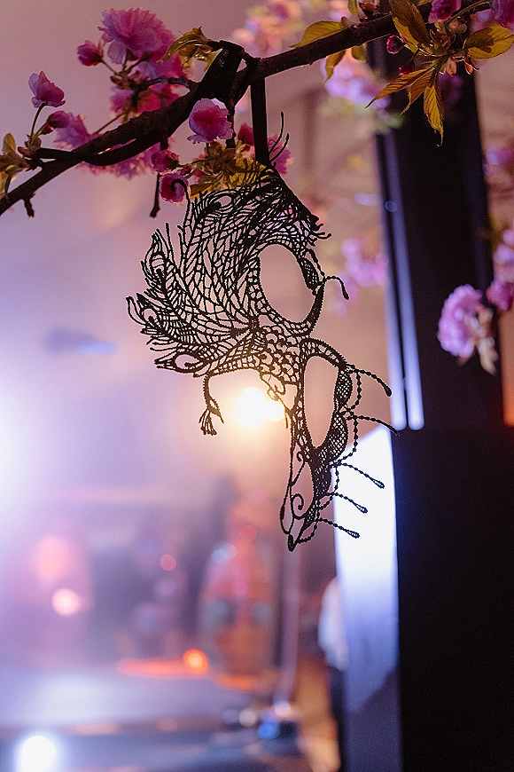 Wedding masquerade mask in black lace with ribbon tie hanging on flowering branches with pink blossoms, against soft indoor bokeh lights