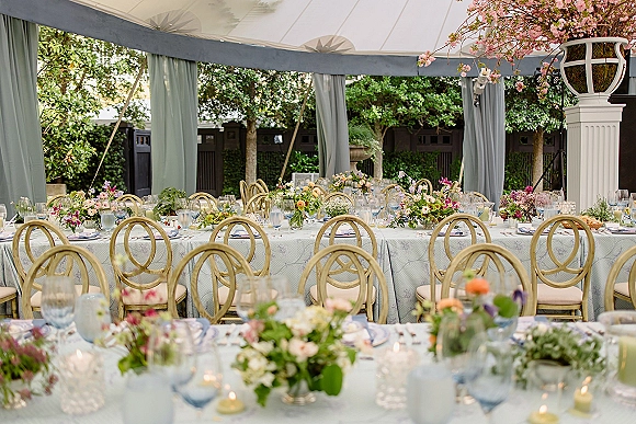 Reception tablescape with an outdoor wedding reception long banquet table set with blue goblets, taper candles, and wildflower centerpieces under a draped canopy in a garden courtyard