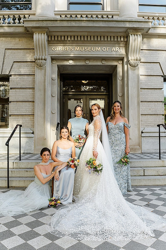 Bridal party portrait with bride centered and bridesmaids in blue dresses holding colorful bouquets on stone building steps by glass doors.