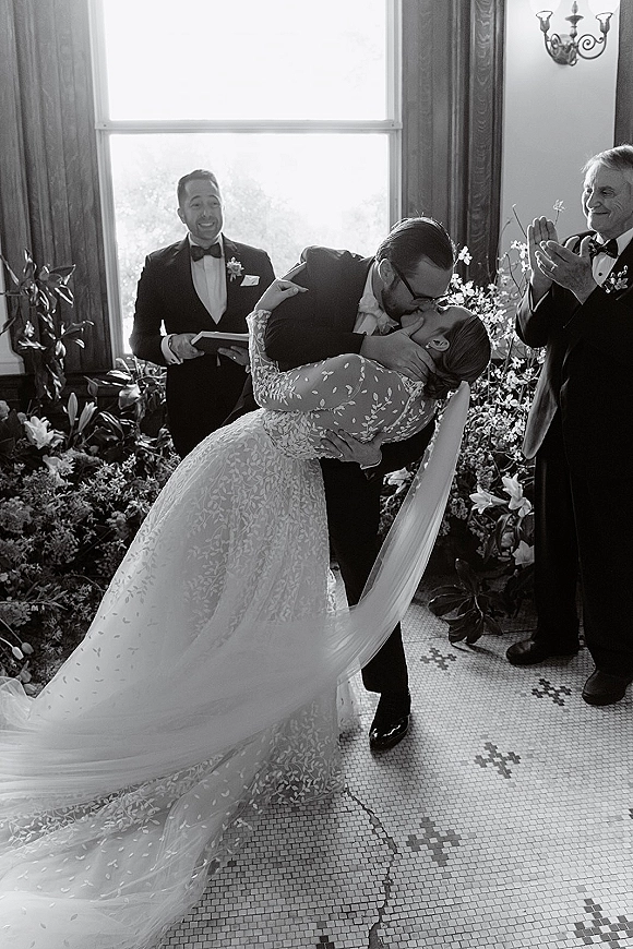 Wedding kiss portrait of bride and groom in a dip, lace dress and long veil, tuxedo with boutonniere, in a window-lit indoor ceremony room