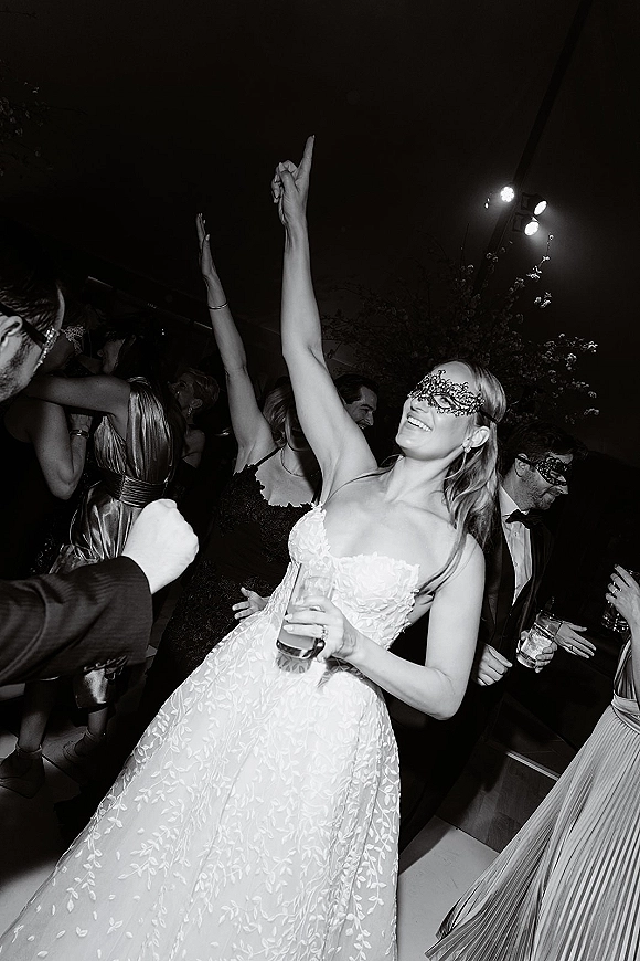 Wedding dance floor packed with guests as the bride dancing at reception in a strapless lace gown, wearing a masquerade mask under stage lighting