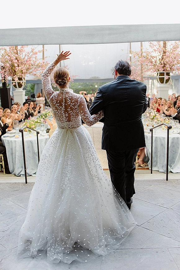 Wedding reception entrance as bride in lace-sleeved tulle gown waves beside groom in black suit, guests cheering by windowed banquet tables with blossoms