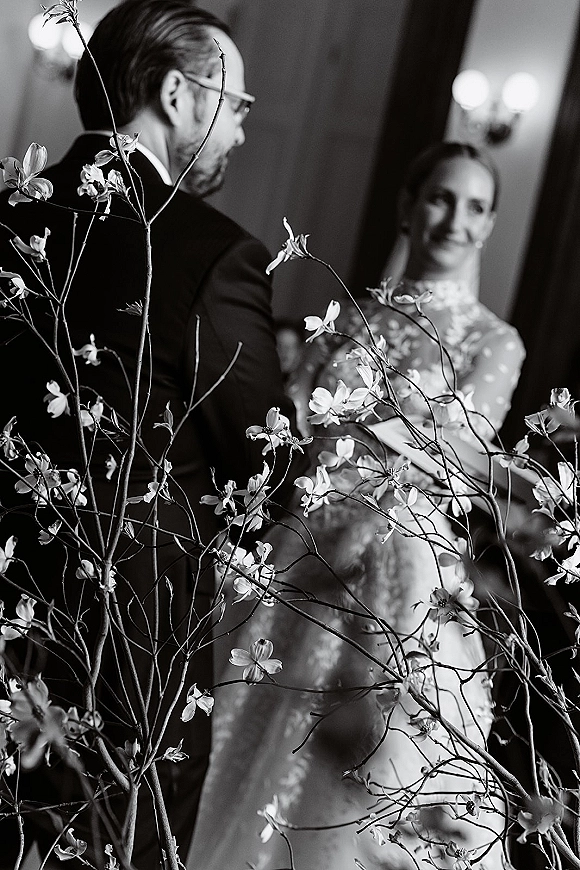 Ceremony moment during wedding vows as bride smiles at groom in glasses, veil and lace gown, framed by floral branches and sconces