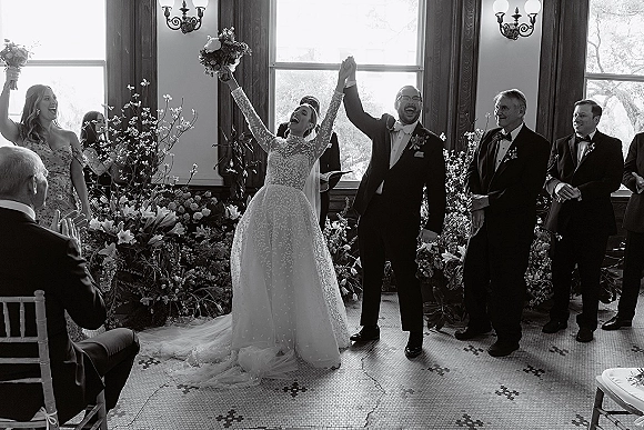 Ceremony moment as bride and groom raise hands, bouquet overhead, with cheering wedding party in bright room with large windows and greenery decor