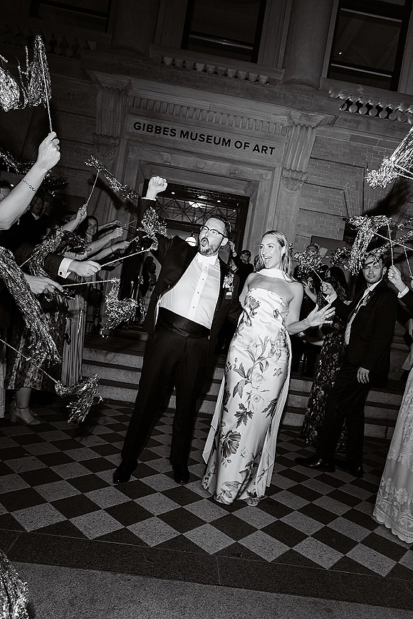 Wedding exit with sparkler send off as bride in strapless floral gown and groom in tux with boutonniere walk past cheering guests at museum steps