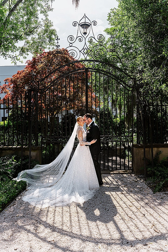 Wedding couple portrait of bride and groom embrace, her long veil and lace sleeves flowing by a wrought iron gate on a garden path