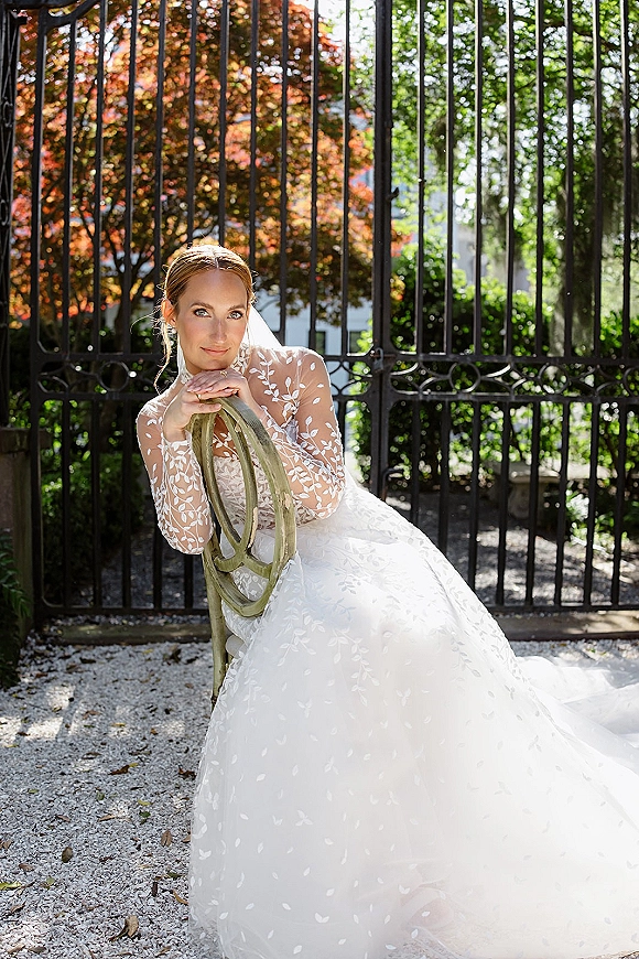 Bridal portrait of a bride in a long sleeve lace gown and veil, leaning on a chair by a sunlit wrought iron gate in a garden path