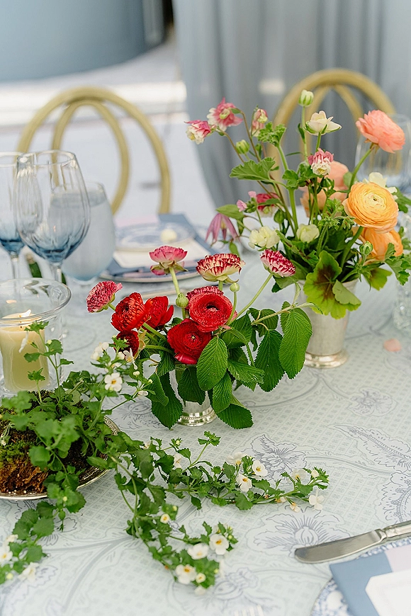 Reception tablescape with wedding table centerpiece of ranunculus blooms and greenery garland, blue goblets, silver flatware, and candles by gray drapery