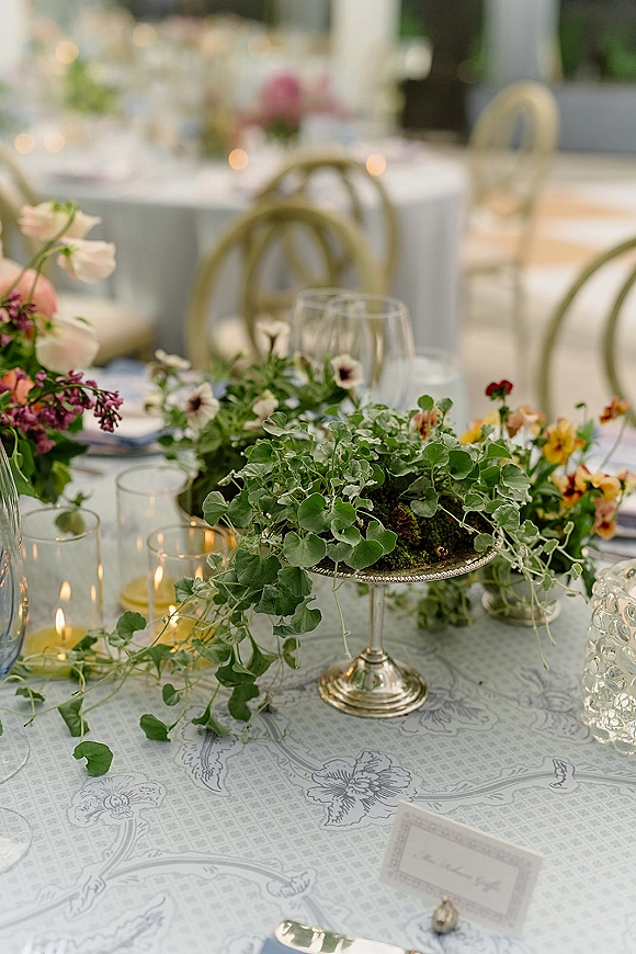 Reception tablescape with wedding table centerpiece of greenery garland, pastel florals, and taper candles on a patterned cloth under a softly lit tent