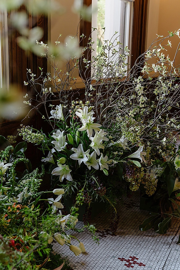 Wedding floral arrangement with white lily wedding flowers and flowering branches, greenery, and bud vases on a tiled floor by an interior window