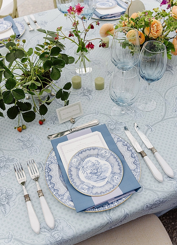 Reception tablescape with blue and white wedding tablescape details—gold rim plates, menus, place cards, floral centerpiece, and small candles