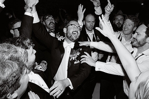 Wedding dance floor packed with groomsmen dance party energy, men in tuxedos and bow ties dancing with hands up in a dark reception crowd