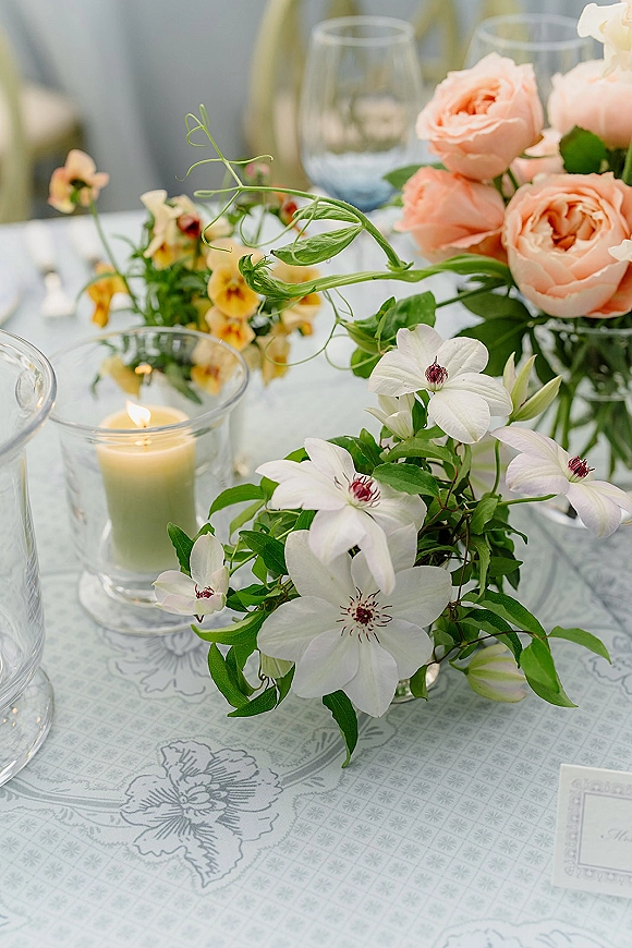 Wedding tablescape with wedding table centerpiece of peach roses, white clematis, greenery and a glass votive candle on patterned tablecloth