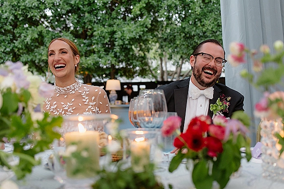 Reception toast moment as bride and groom laughing at the head table, surrounded by candles and floral centerpieces under draped trees