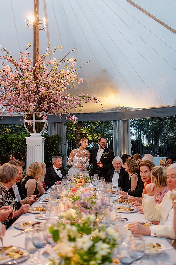 Wedding reception toast as bride and groom speech with raised cocktail glasses at a long banquet table under string-lit white tent