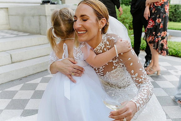 Bride hugging child in a long sleeve lace wedding dress, veil, and champagne coupe, smiling on stone steps with guests nearby