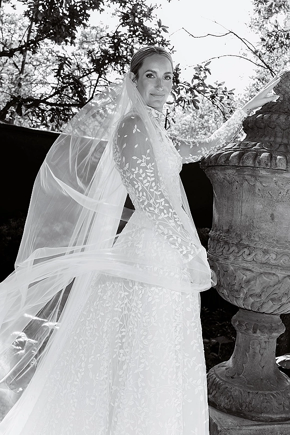 Bridal portrait of a bride in a long sleeve lace gown with cathedral veil, side profile by a stone urn amid garden trees and hedge