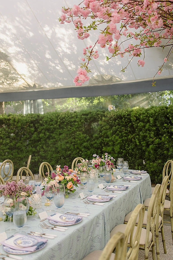 Reception tablescape with outdoor wedding tablescape accents, blue stemware and floral centerpieces on a long banquet table under a tent canopy