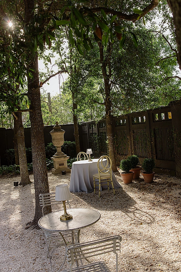 Wedding lounge area with outdoor wedding lounge bistro tables, white tablecloths and table lamps under a tree canopy on gravel in a garden setting