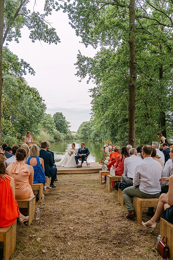 Outdoor wedding ceremony with riverside wedding ceremony seating on wood benches facing a wooden stage with floral arrangement by the river