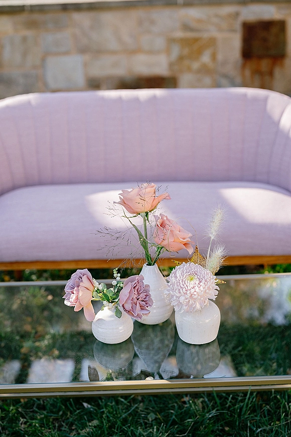 Wedding floral arrangement in white ceramic bud vases with blush and mauve roses on a glass coffee table beside a blush sofa outdoors