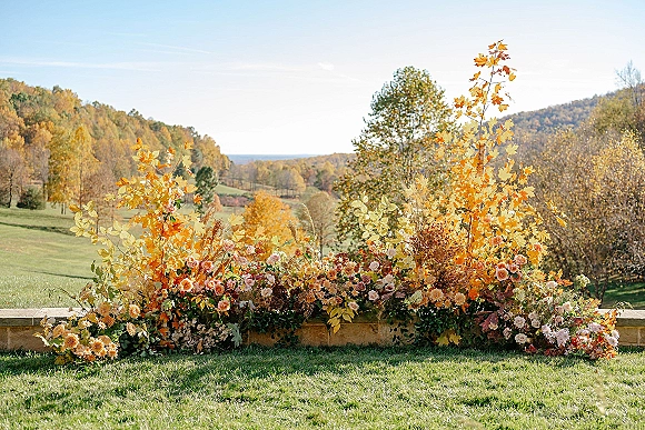 Wedding ceremony florals in a ground floral ceremony arrangement with roses, dahlias, and fall foliage on a lawn by rolling hills under blue sky