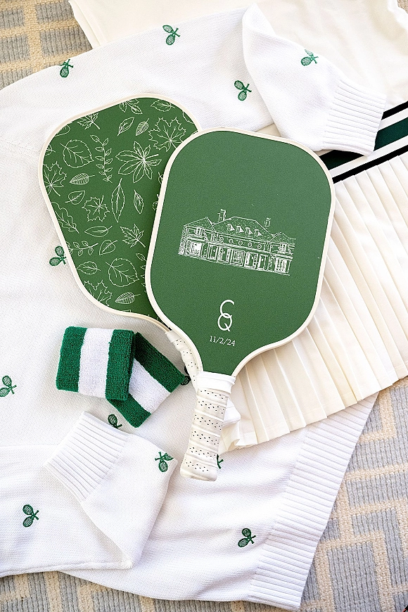 Wedding welcome bags with monogram paddle fans, striped wristbands, and embroidered tennis socks arranged on white knit fabric and a beige rug