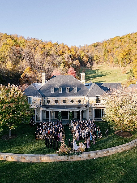 Outdoor wedding ceremony with aerial wedding ceremony view of guests seated by aisle florals on a lawn before a stone manor house under blue sky